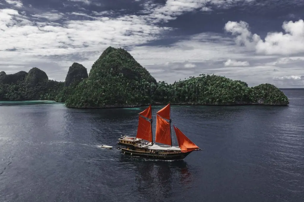 The Phinisi boat Calico Jack with its striking red sails contrasting against a tropical island backdrop under a partly cloudy sky 
