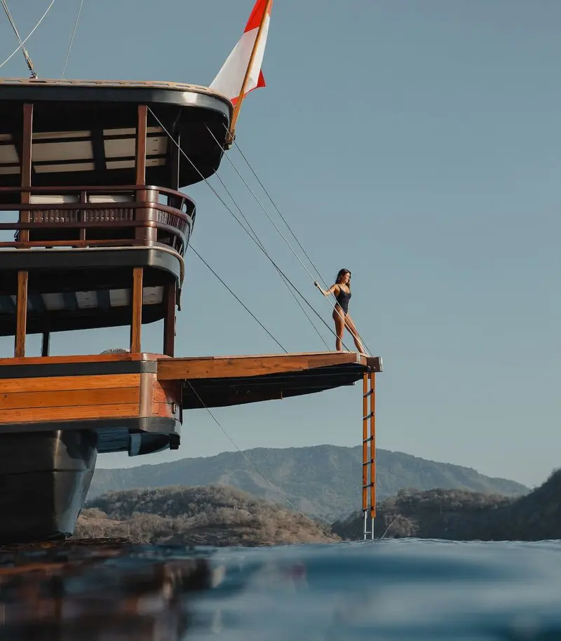 A beautiful women in black swimsuit stands on the stern dive platform of the yacht Dunia Baru, with tropical islands and blue sky behind.