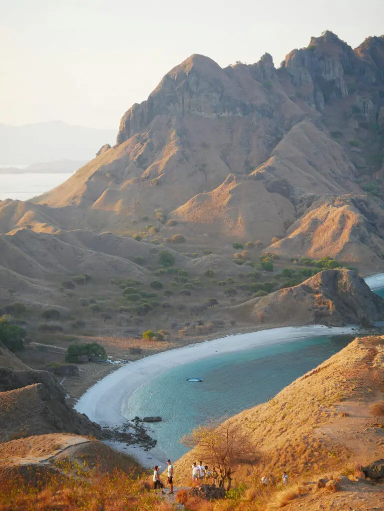 The dry, arid and mountainous landscape of Komodo meets the white sand beaches and bright blue waters of the ocean, while a several people take in the views.