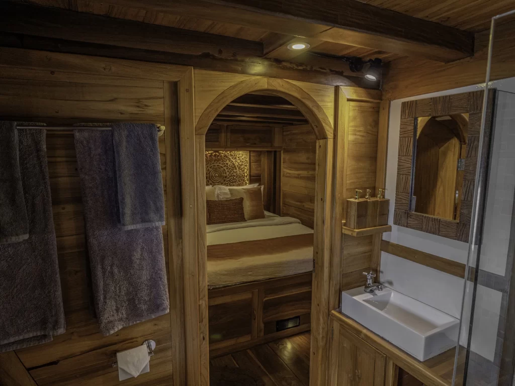 A cozy wooden cabin bathroom, reminiscent of a charter yacht in Indonesia, features a white sink, mirror, glass shower, and shelves. Through an arched doorway, a neatly made bed with pillows and rustic wood walls is visible. Two towels hang on a rack.