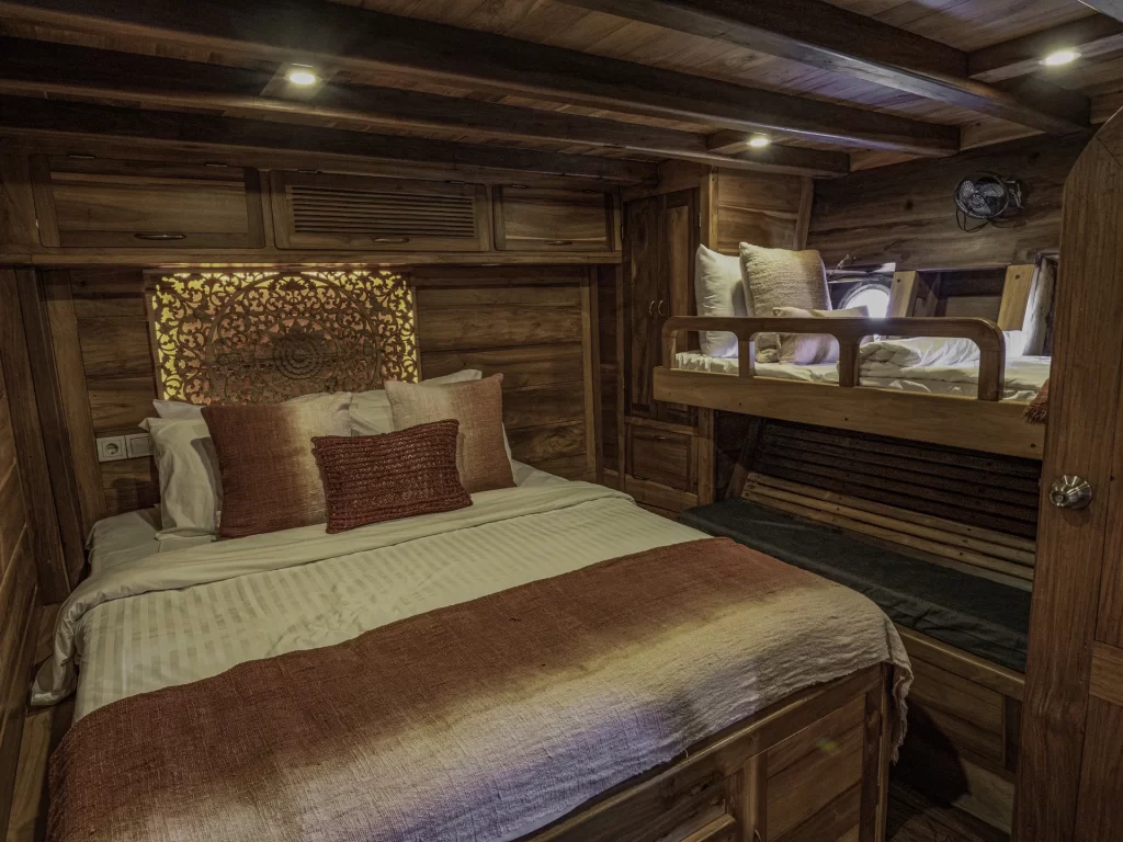 Cozy wooden cabin bedroom with a double bed and a bunk bed, reminiscent of an Indonesia charter yacht. The beds have white linens with brownish-red accents, while warm lighting and intricate headboard detailing create a welcoming atmosphere.