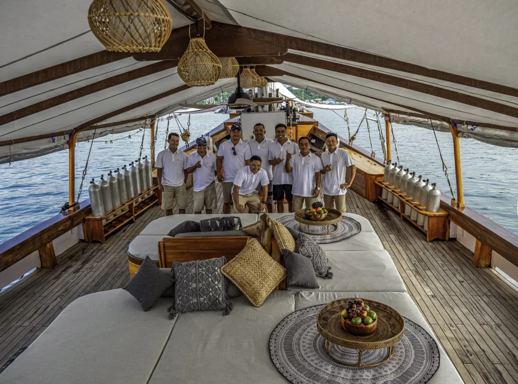 The crew of eight people on Anne Bonny in white shirts and shorts pose and smile on the deck of a wooden boat with cushioned seating, decorative pillows, and fruit bowls, with water and land visible in the background.