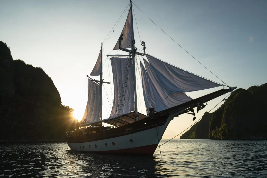 A charter yacht with large white sails glides on calm water at sunset in Indonesia, with cliffs and trees silhouetted in the background. The sun is partially visible behind the boat, creating a warm, serene atmosphere.